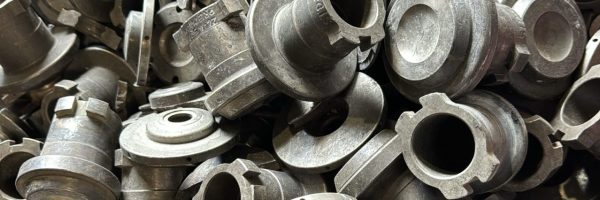 Pila de piezas metálicas industriales para reciclaje: componentes cilíndricos con bridas, ranuras y orificios, superficies grises con desgaste visible.